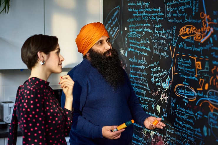 Un homme portant un turban orange parle avec une collègue devant un tableau noir