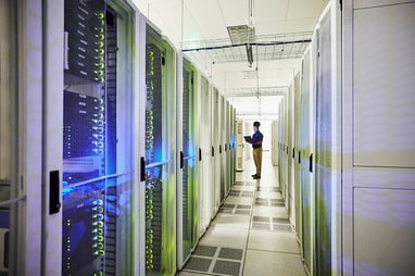A man stands while working on a laptop in a corridor of servers. 