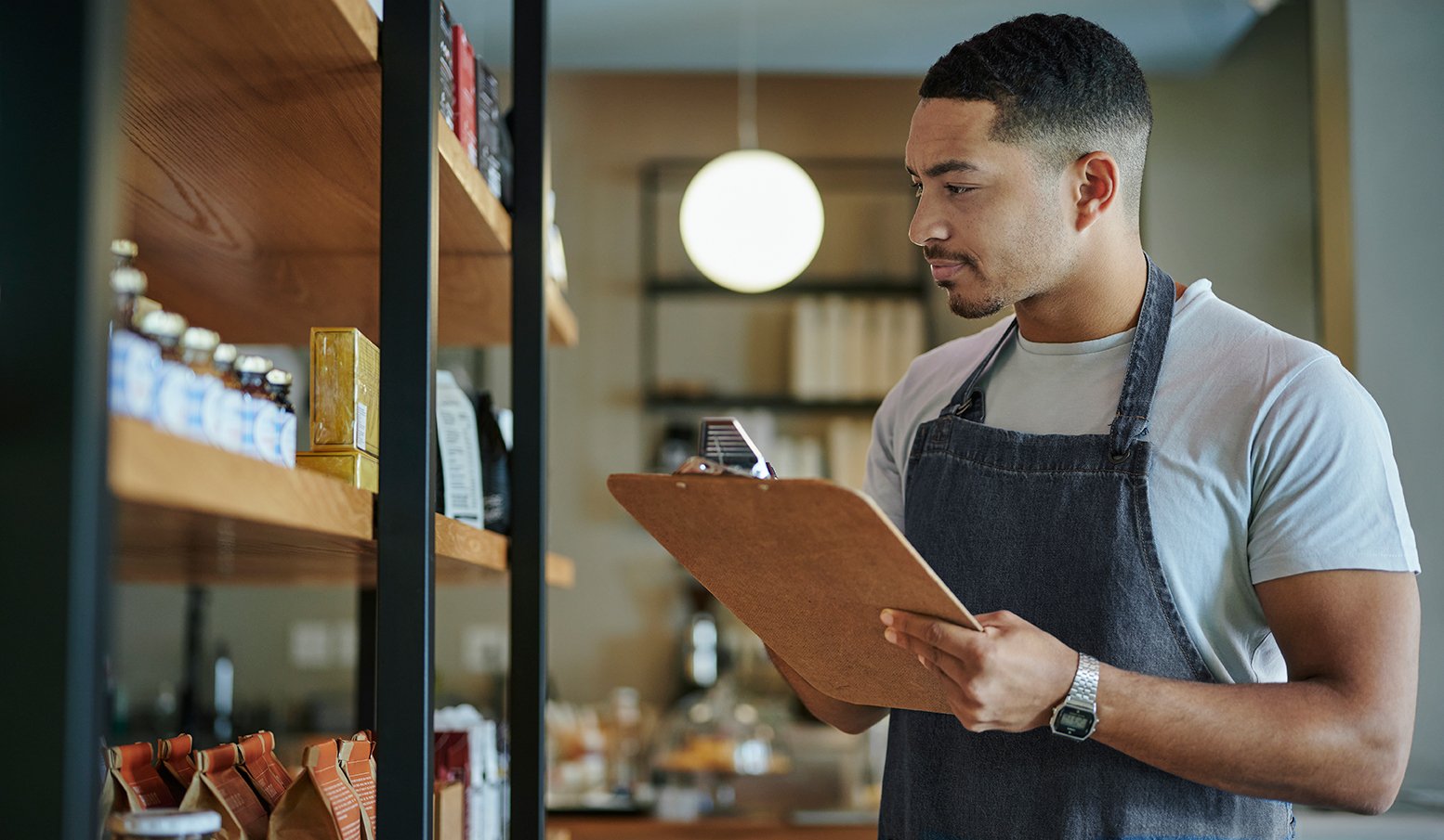 A young business owner holds a clipboard while looking at inventory on a shelf.